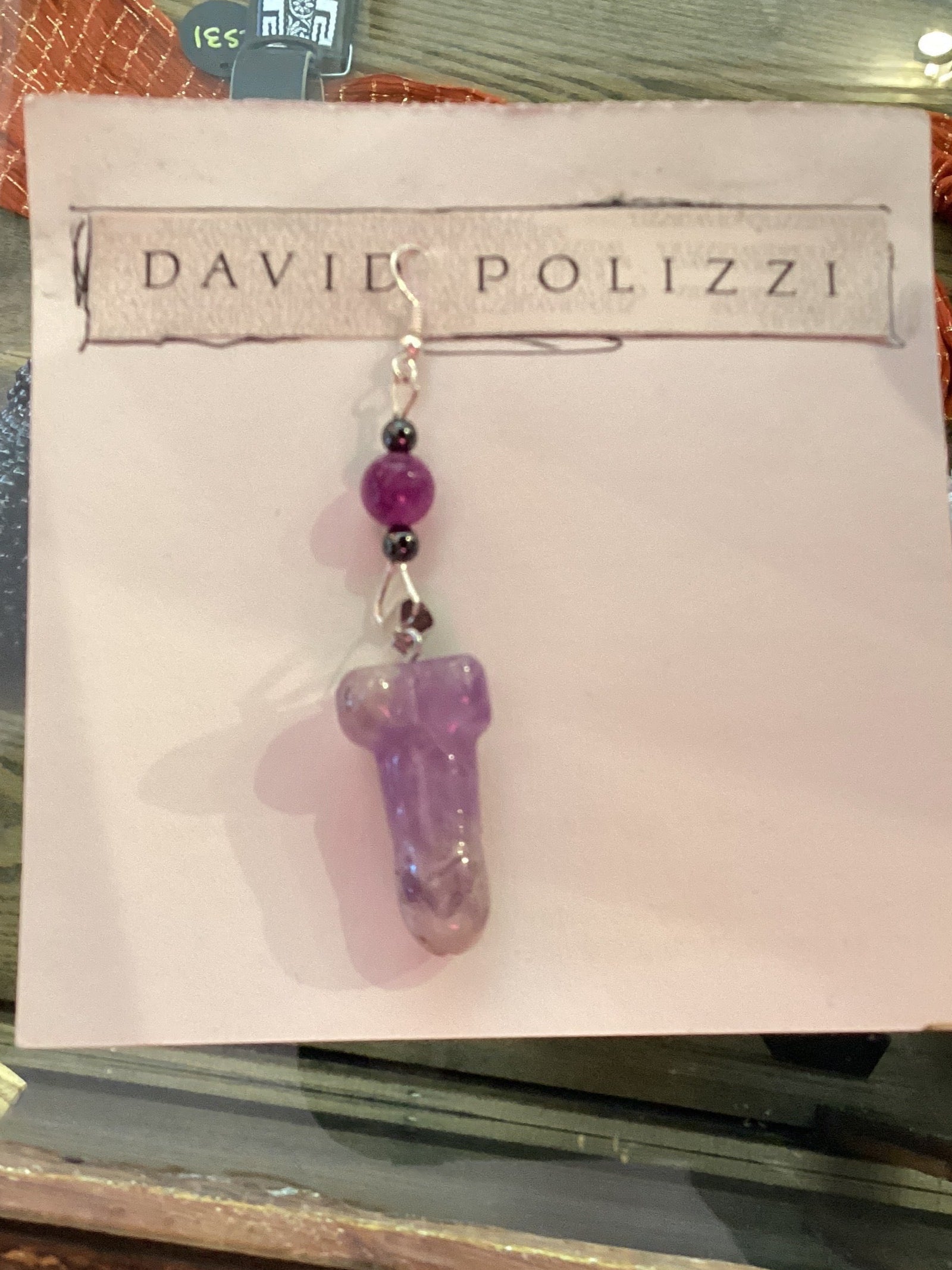 This image displays a pair of intricately designed earrings, titled "David Polizzi Stone Penis Earrings." The earrings are crafted with a smooth, purple stone shaped in a playful, phallic form, set in simple, elegant settings with small silver hooks.